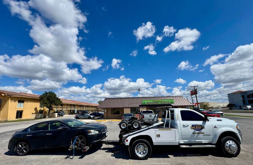 A white tow truck towing a dark sedan in a paved parking lot under a bright, cloudy blue sky.