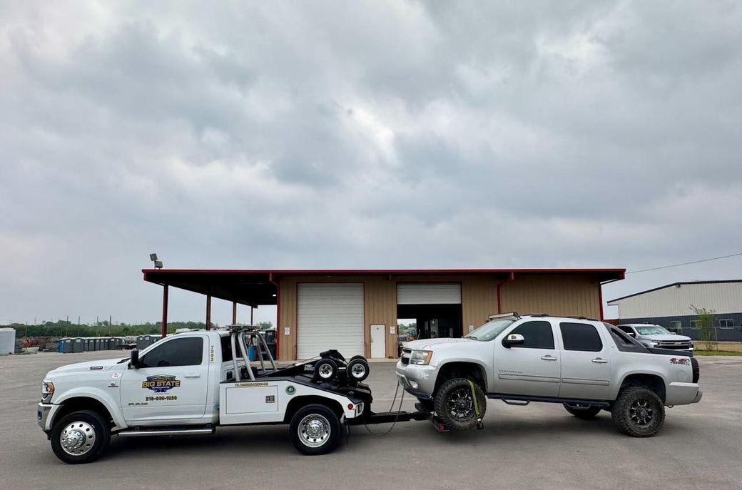 A white tow truck towing a silver pickup truck in front of a metal warehouse building on a cloudy day.