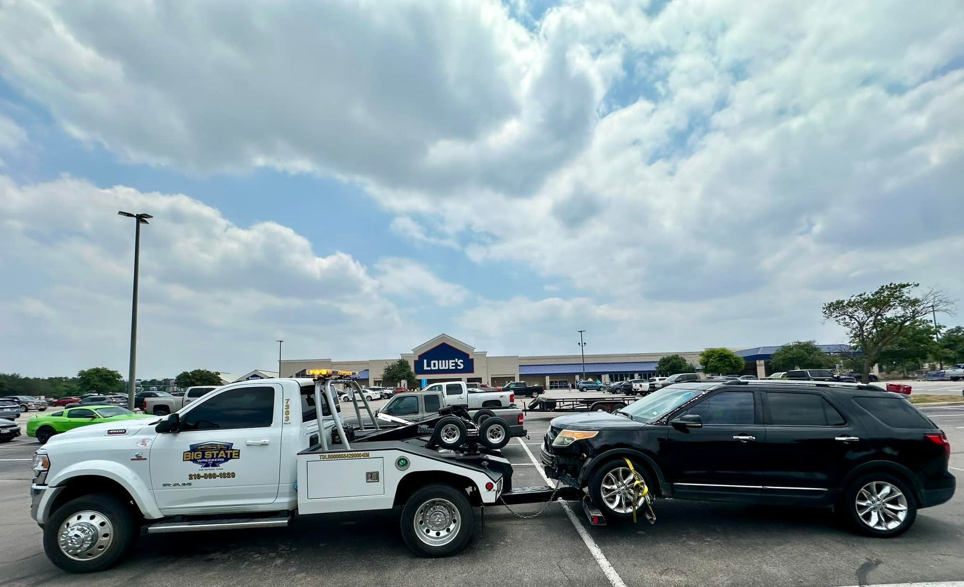 A white tow truck is towing a black SUV in a parking lot on a cloudy day, with a Lowes store in the background.