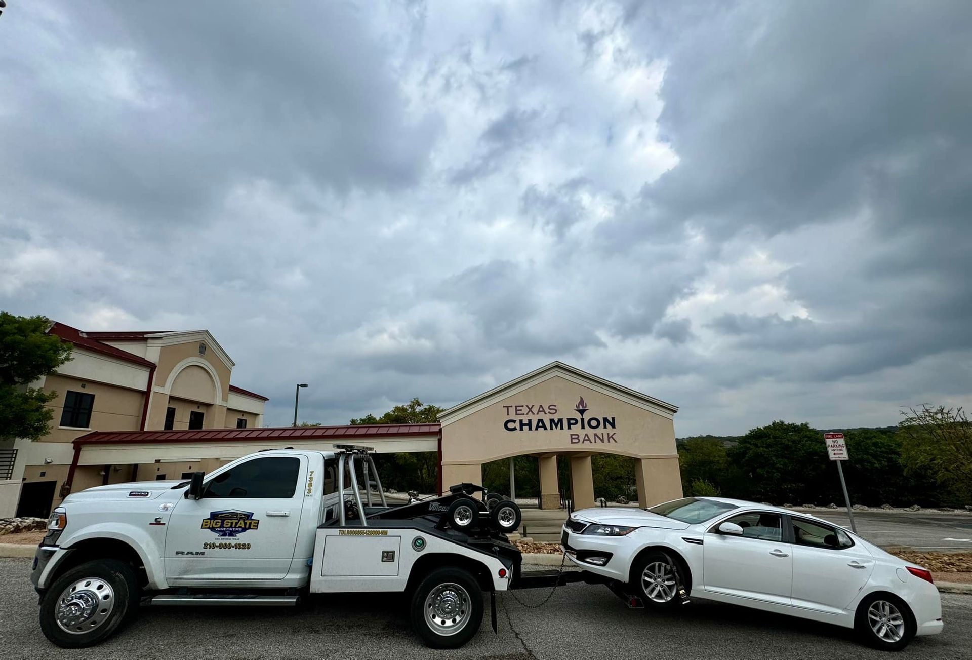 A white tow truck tows a white sedan in front of a building labeled Texas Champion Bank under a cloudy sky.
