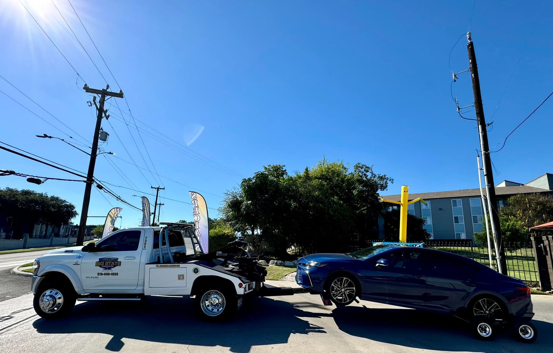 A white tow truck towing a dark blue car on a sunny day with a blue sky and utility poles in the background.