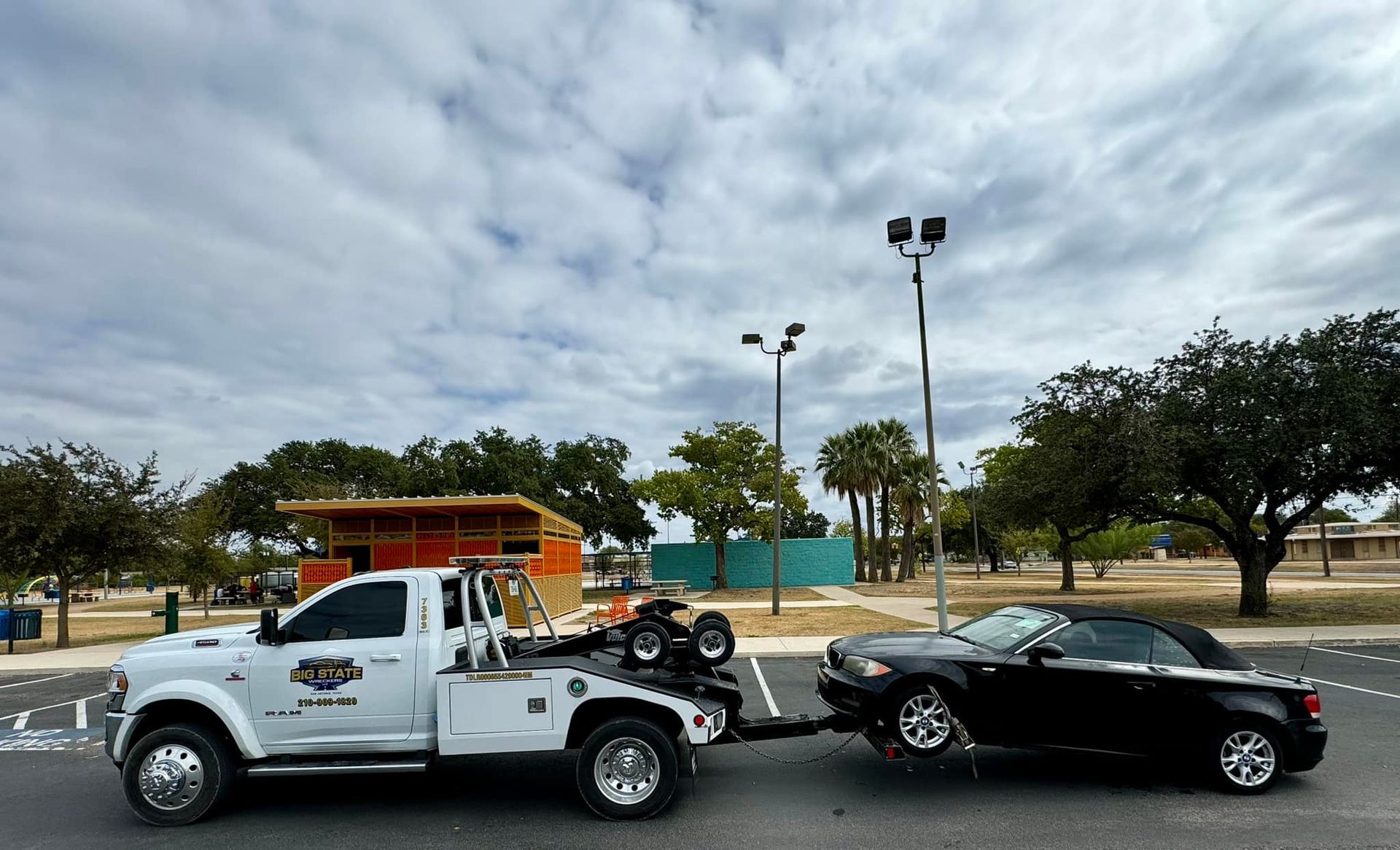 A white tow truck towing a black convertible car in a parking lot under a cloudy sky.