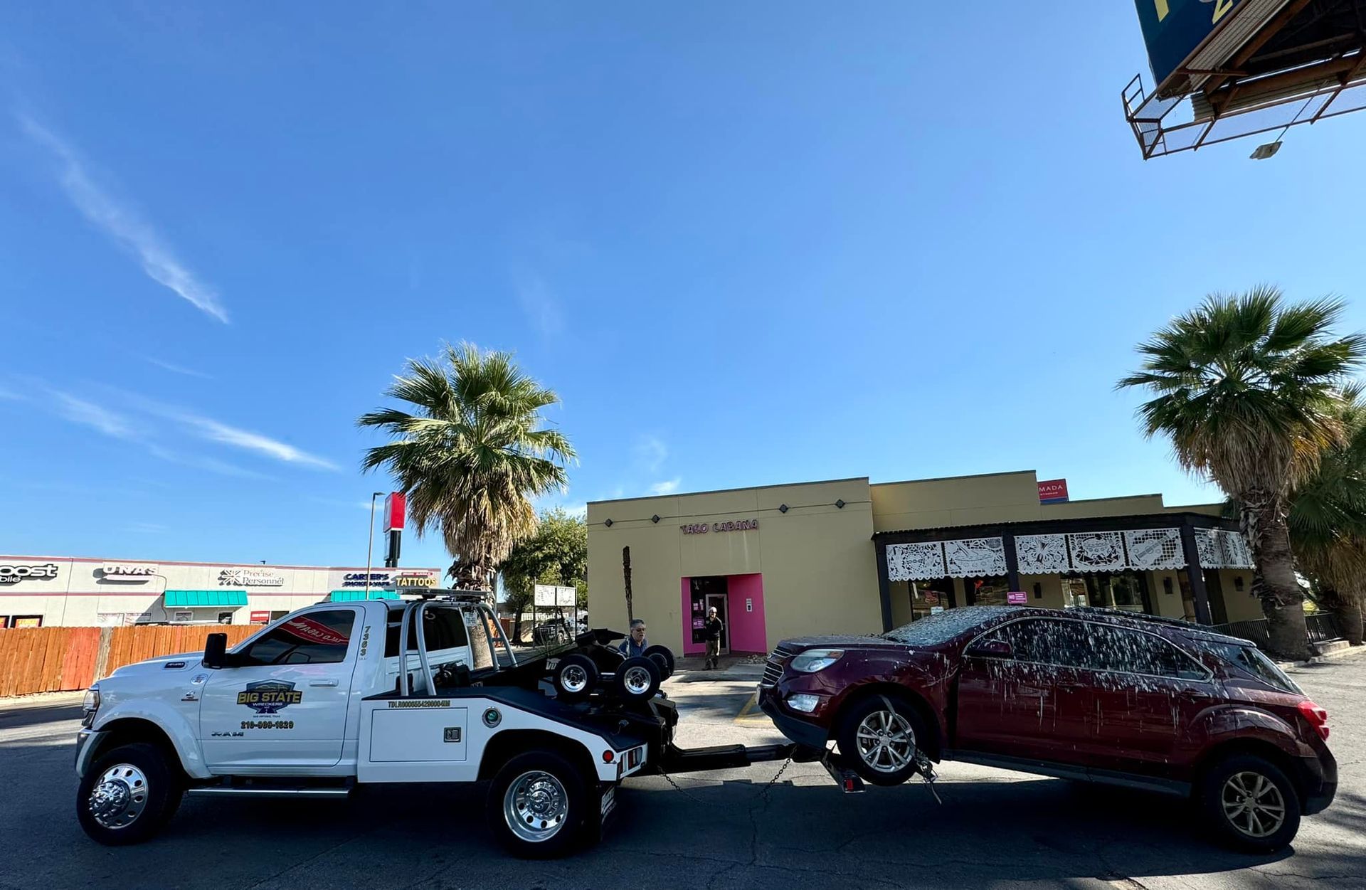 A white tow truck is hitched to a maroon SUV, towing it across a parking lot on a sunny day with palm trees in the background.