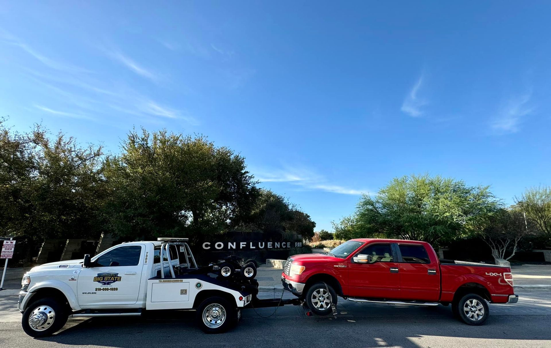 A white tow truck towing a red pickup truck against a blue sky with trees and a building in the background.