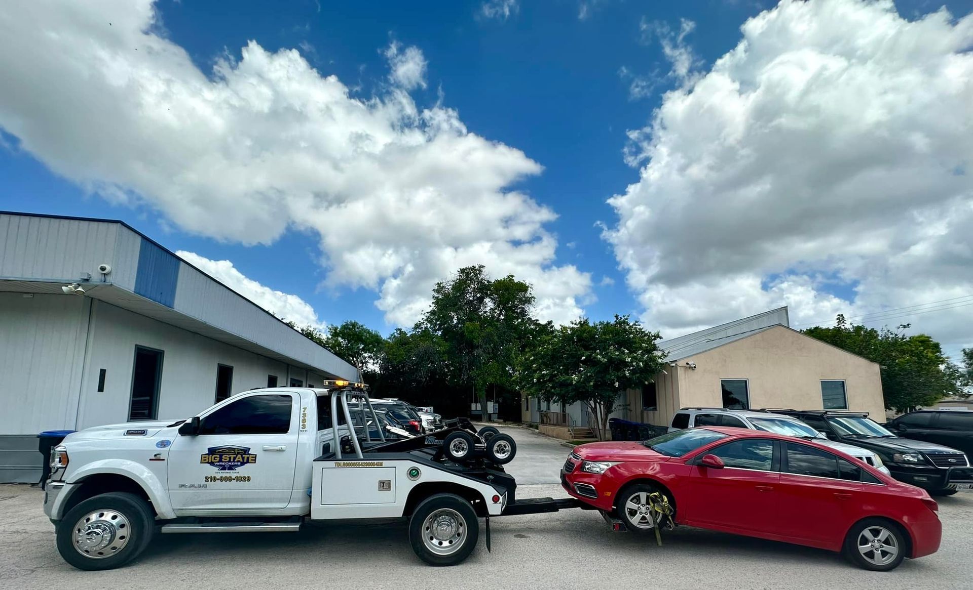 A white tow truck is towing a red sedan in a gravel parking lot under a bright blue sky with large clouds.