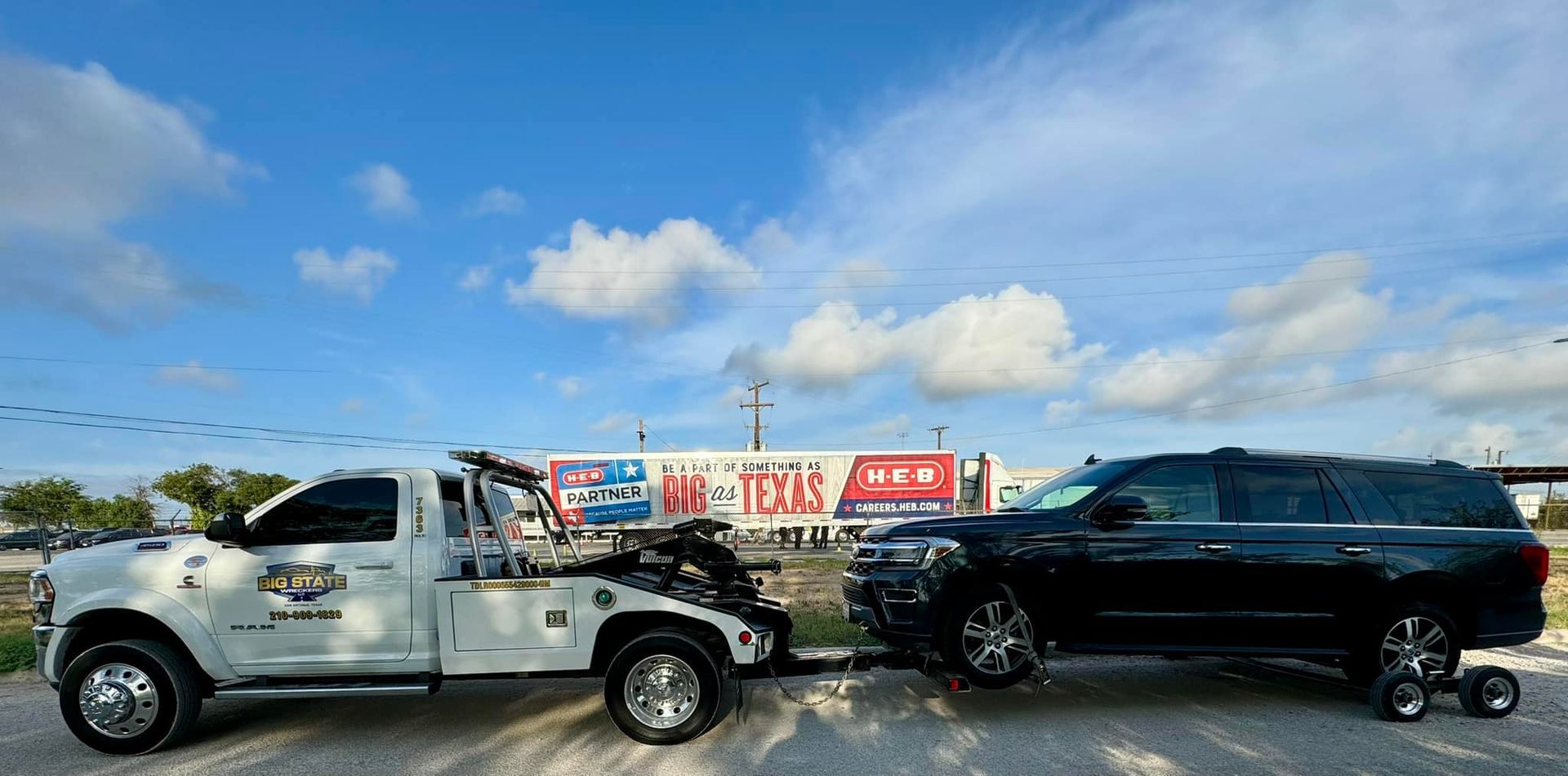 A white tow truck is towing a black SUV on a gravel lot under a clear blue sky with white clouds.