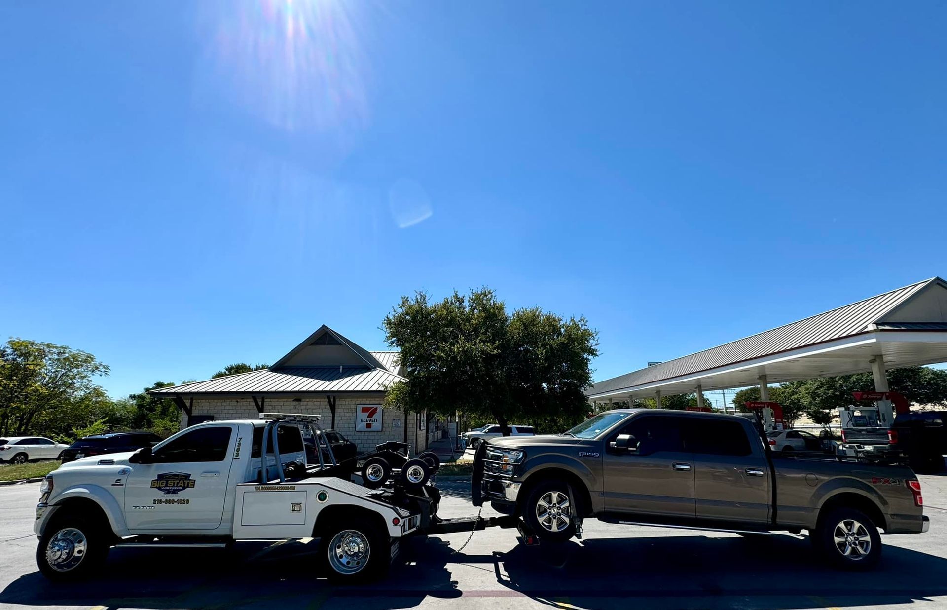 A white tow truck is towing a grey pickup truck in a parking lot on a bright, sunny day.