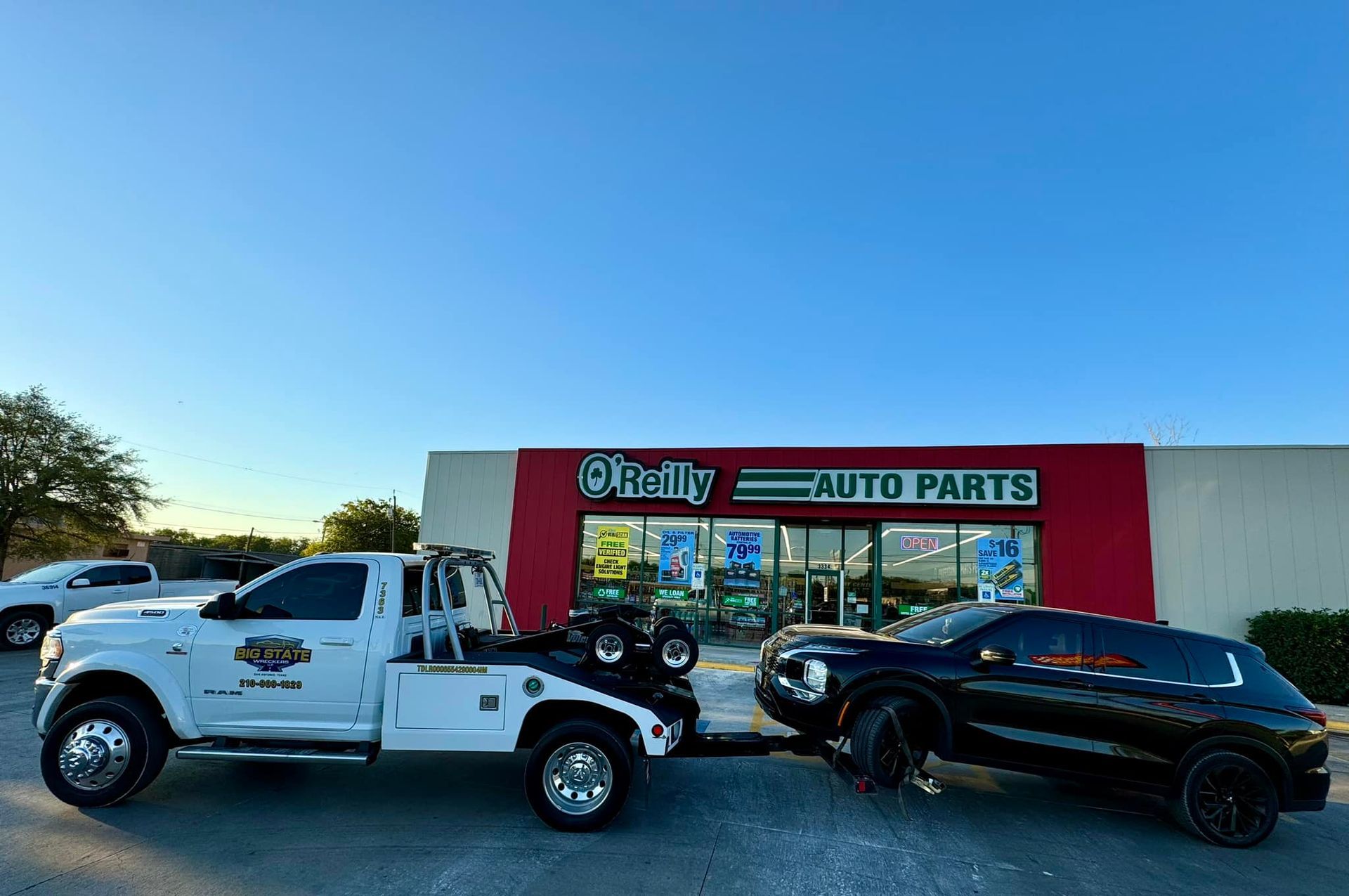 A white tow truck towing a black SUV in front of an O'Reilly Auto Parts store under a clear blue sky.