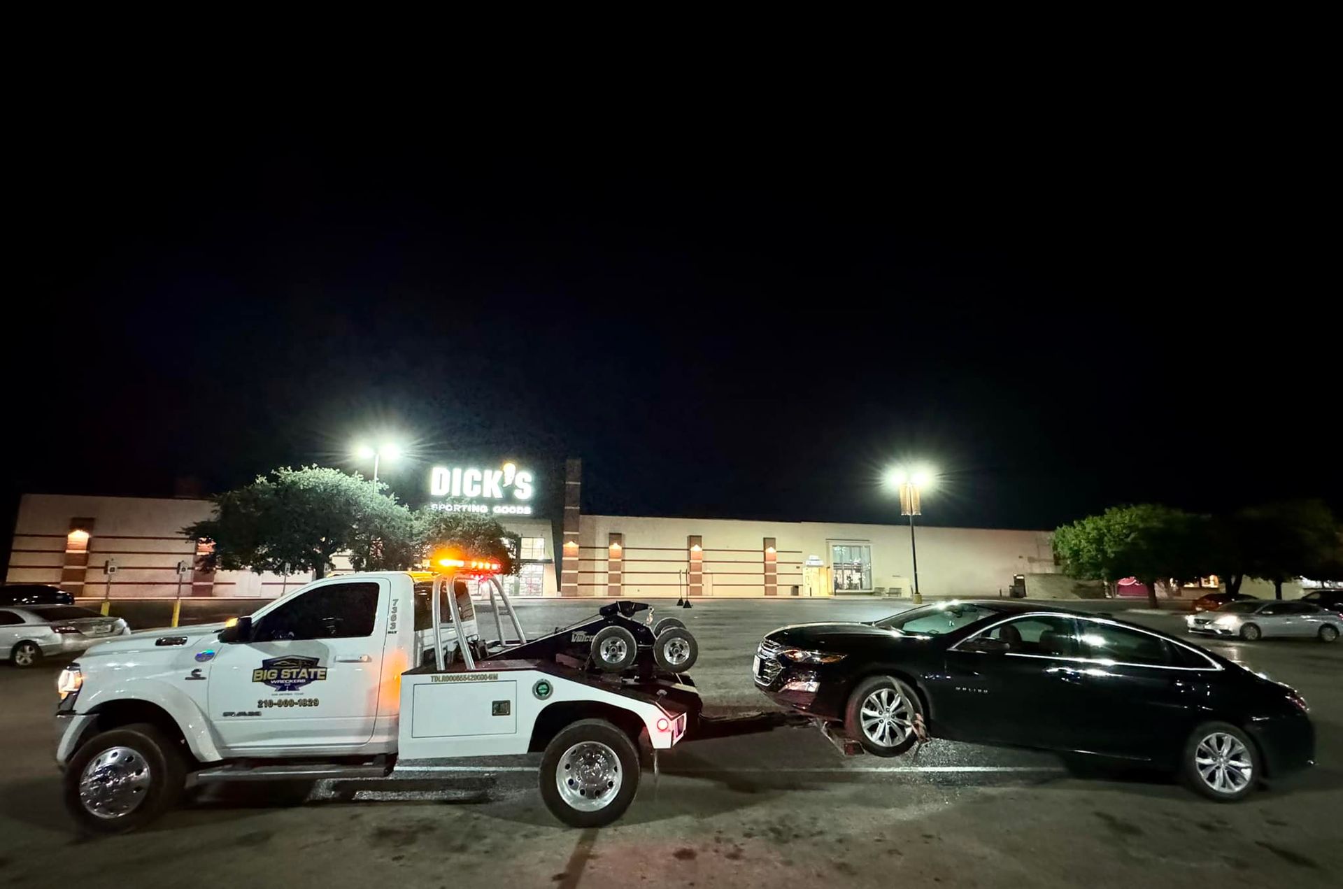 A white tow truck tows a black sedan in a shopping center parking lot at night, with a Dick's Sporting Goods store behind.