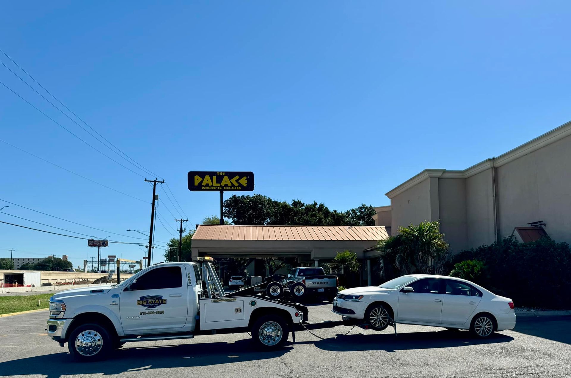 A white tow truck is towing a white sedan in a parking lot in front of a building with a 