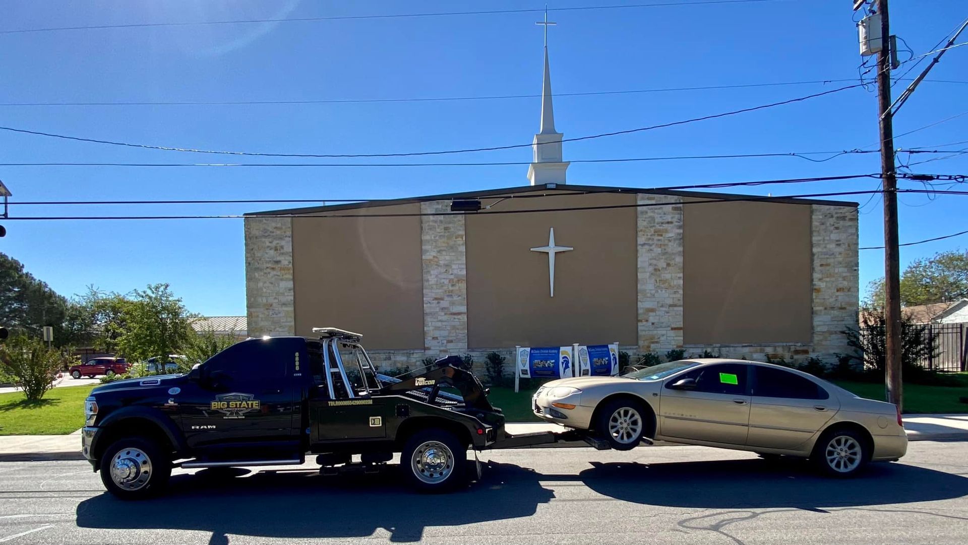 A black tow truck towing a gold sedan on a sunny day in front of a tan church with a steeple and a cross.