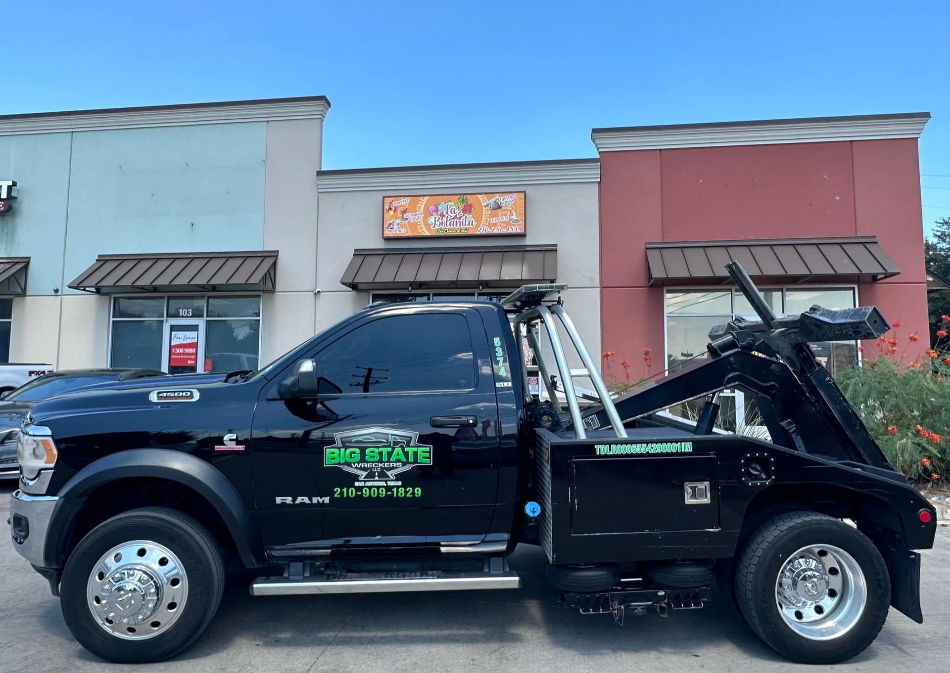 A black tow truck with green logo text is parked in front of a commercial building with light and reddish-brown walls.