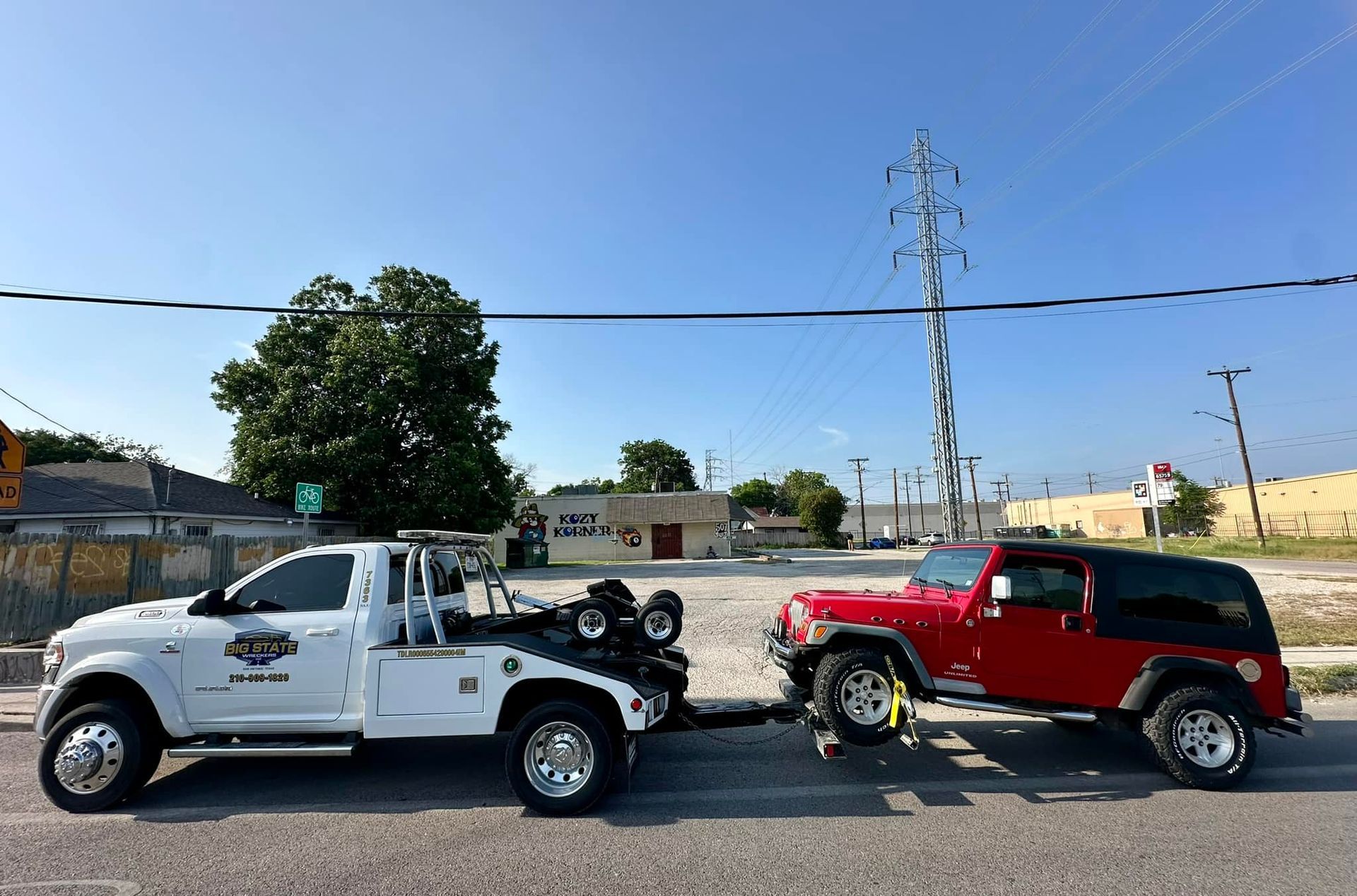 A white tow truck towing a red Jeep Wrangler on a sunny day in a paved lot.