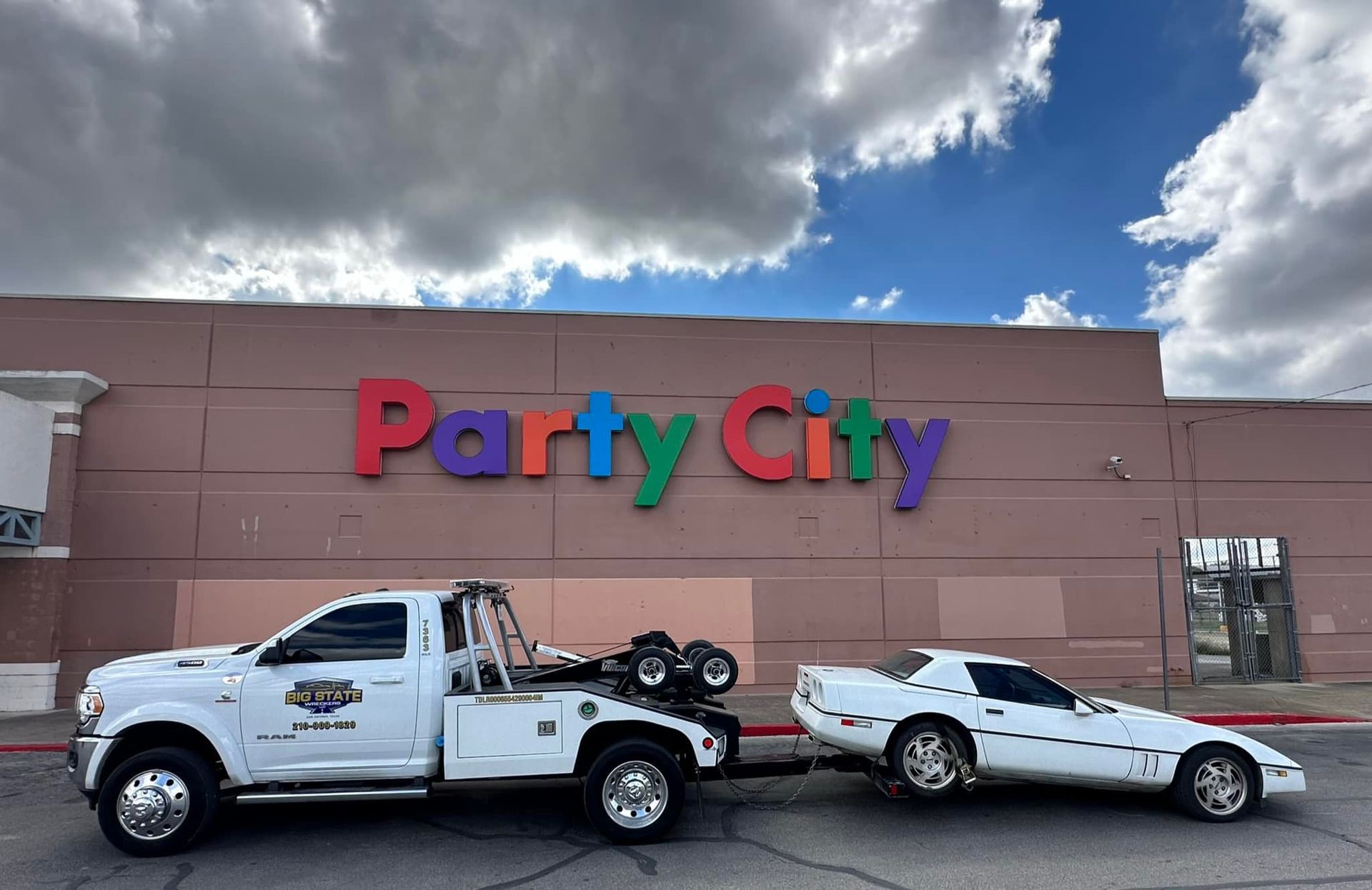A white tow truck is towing a white sports car in a parking lot in front of a Party City store.