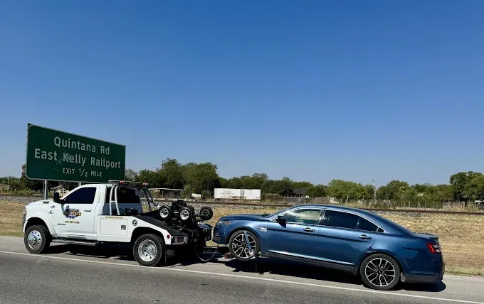 A white tow truck is towing a blue sedan on the side of a highway next to a sign for Quintana Rd and East Kelly Railport.