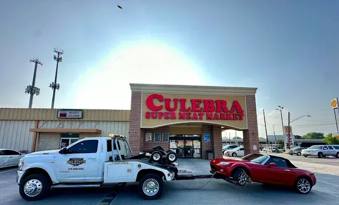 A white tow truck towing a red convertible sports car in front of a Culebra Super Meat Market building.