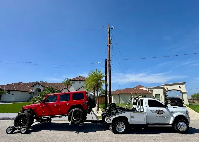 A white tow truck is towing a red Jeep Wrangler on a residential street under a clear blue sky.