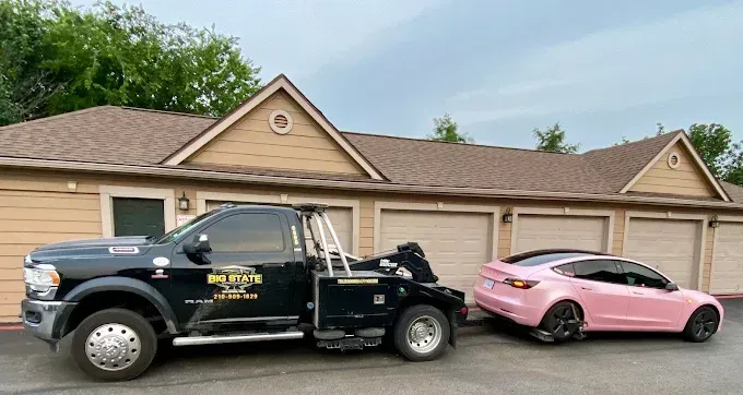 A black tow truck hooks up a light pink Tesla parked in front of a residential building with tan garage doors.