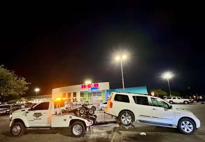 A tow truck in a parking lot at night, towing a white SUV in front of an H-E-B grocery store.
