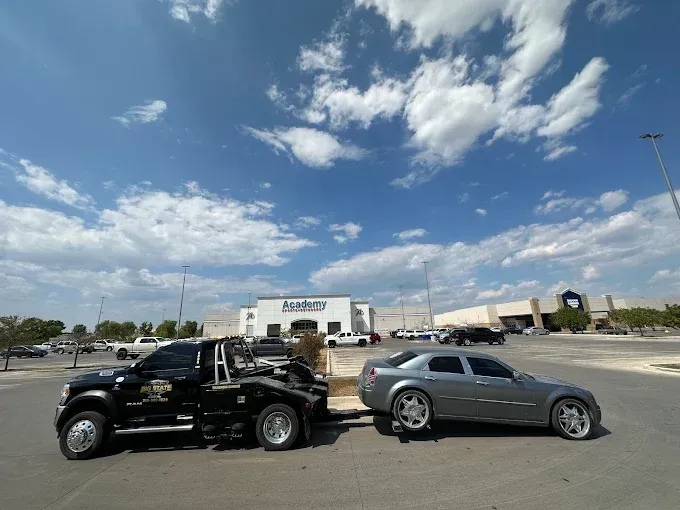 A black tow truck towing a silver Chrysler 300 in a parking lot on a sunny day with a store in the background.