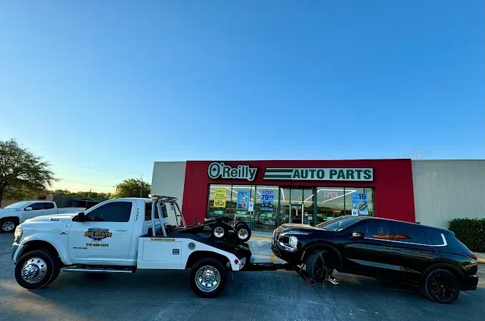 A white tow truck is towing a black SUV in front of an O'Reilly Auto Parts store under a clear blue sky.
