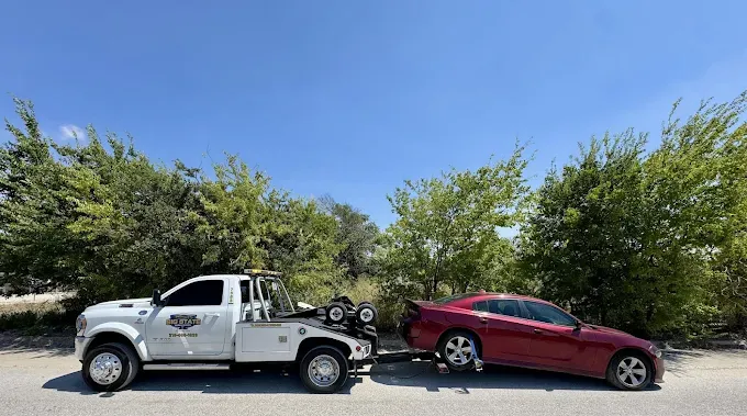 A white tow truck towing a red sedan along a road lined with trees on a sunny day.