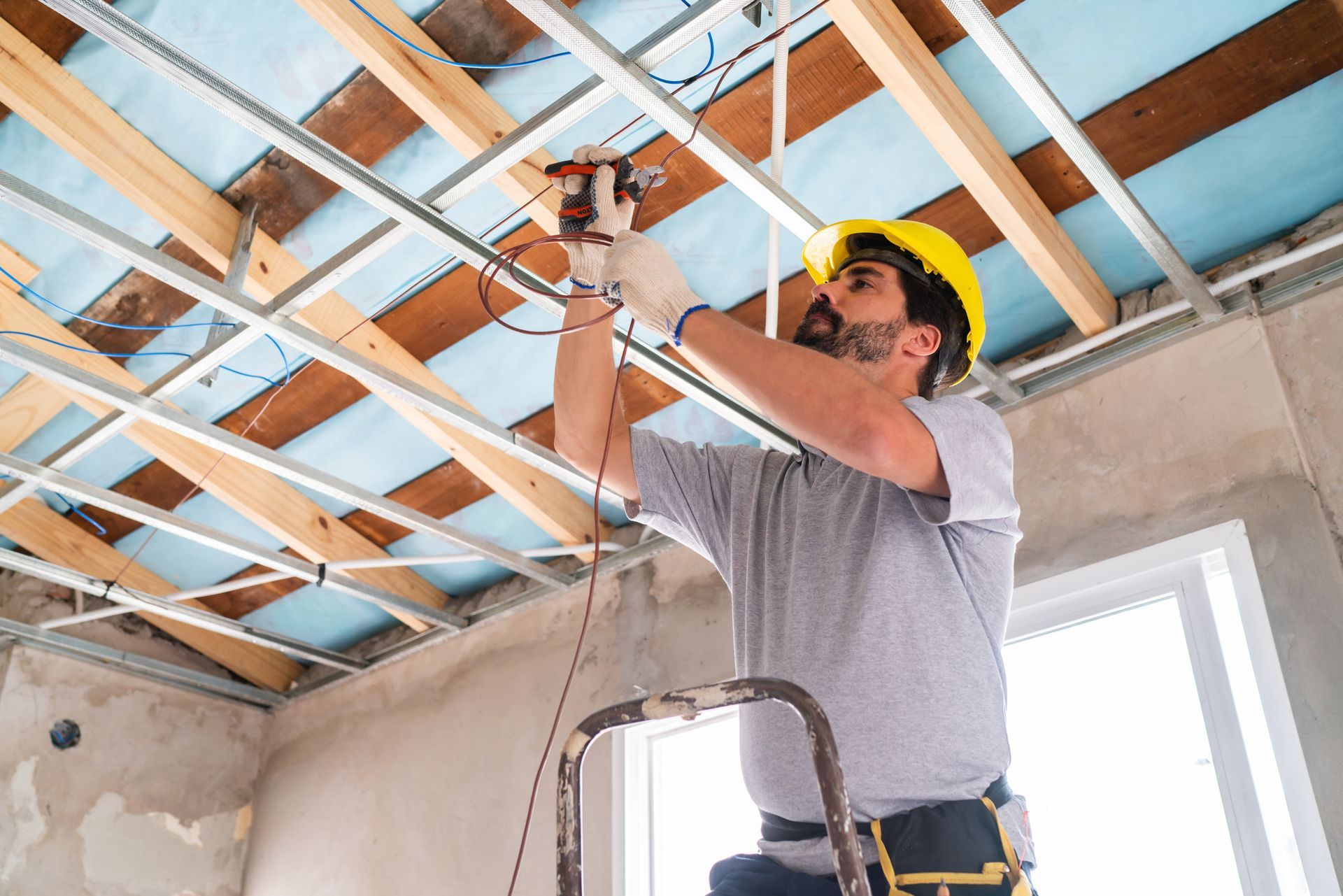 Electrician in yellow hard hat wiring ceiling, standing on ladder.
