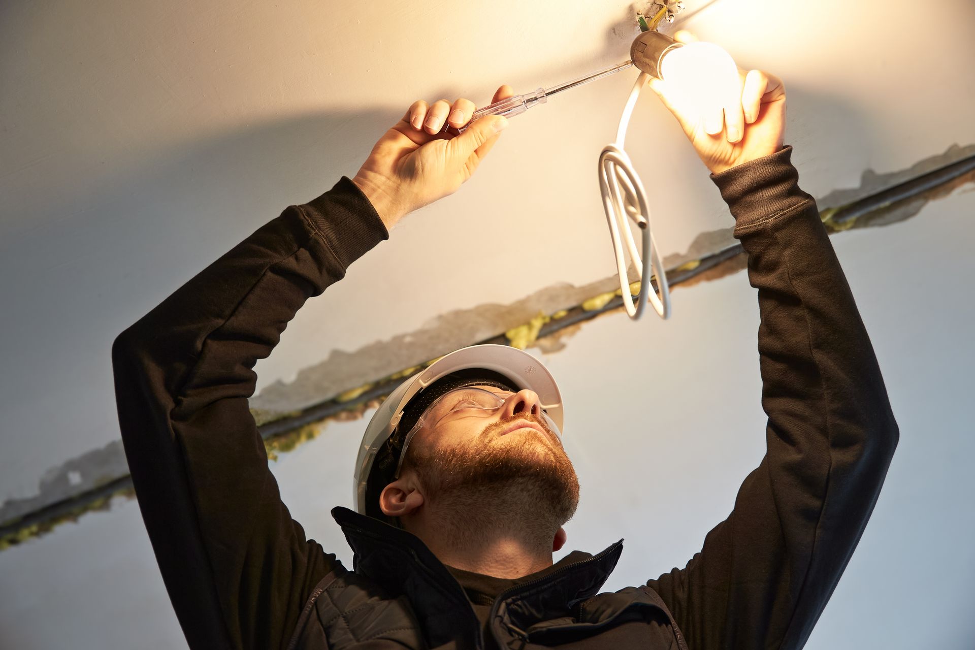 Electrician fixing light fixture, wearing hard hat, arms raised, indoors.