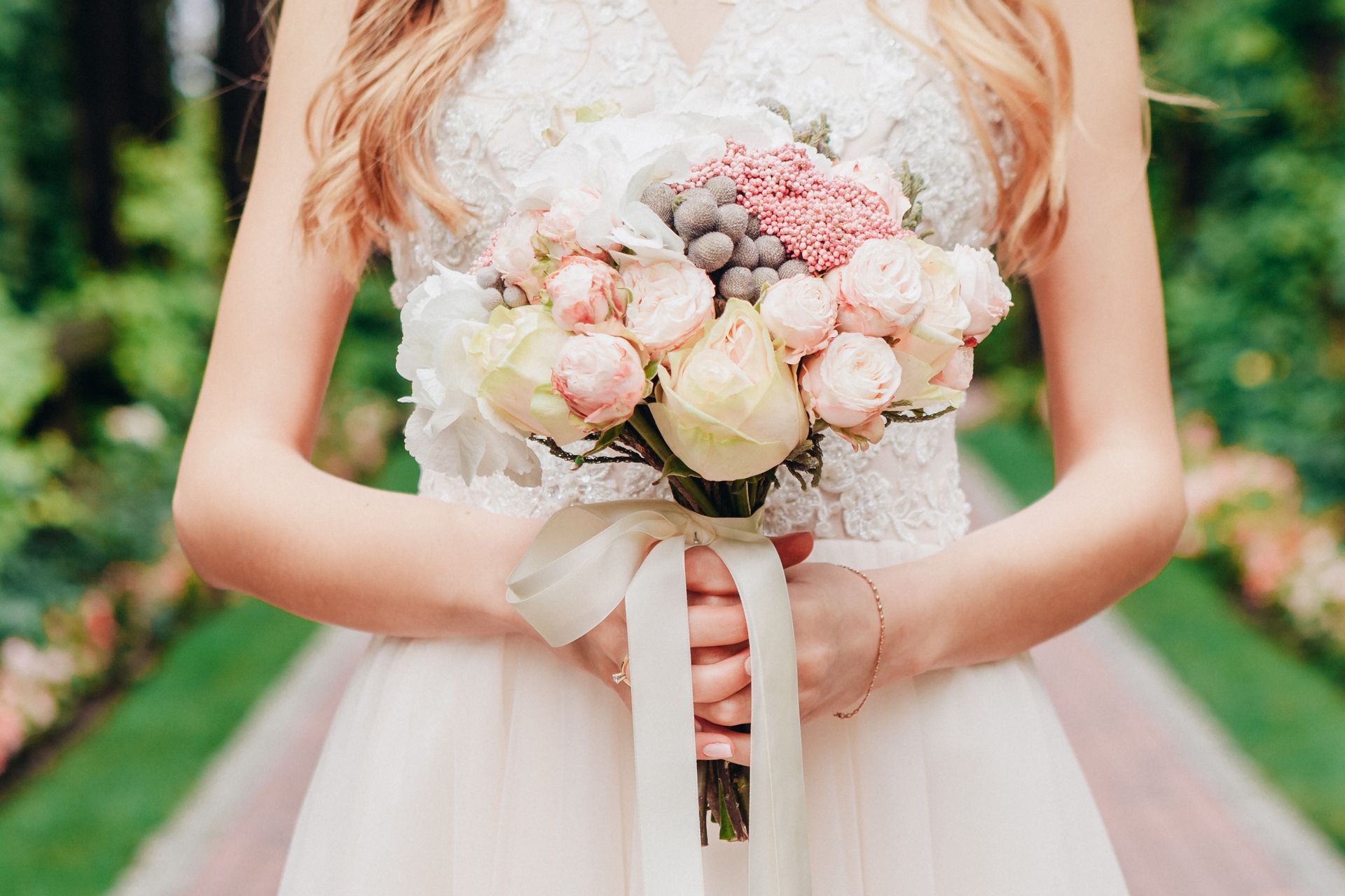 Bride in white dress holding a pastel bouquet with cream ribbon, standing outdoors.