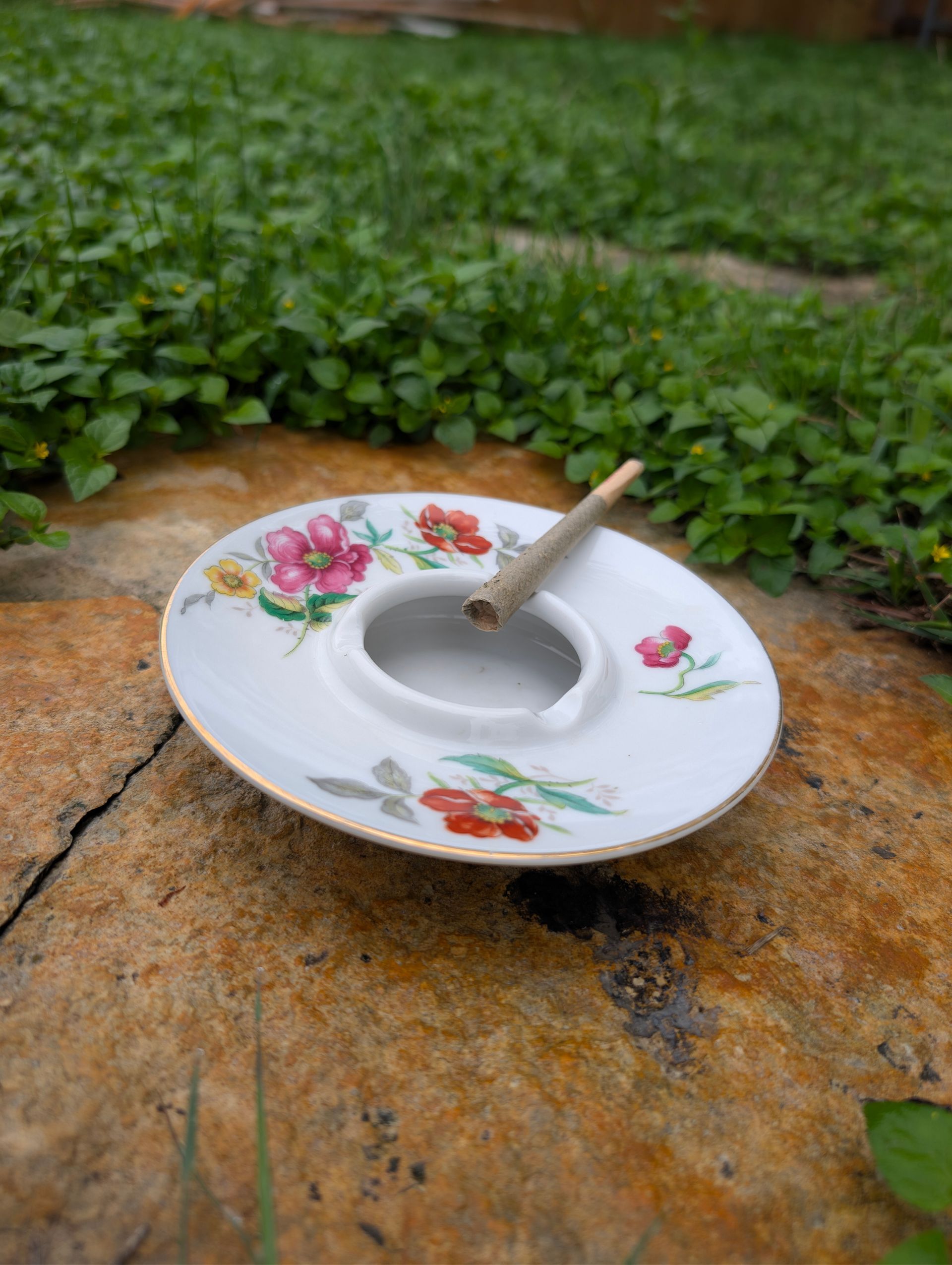 Floral porcelain saucer on a rock with a small spoon, beside green ground cover plants.