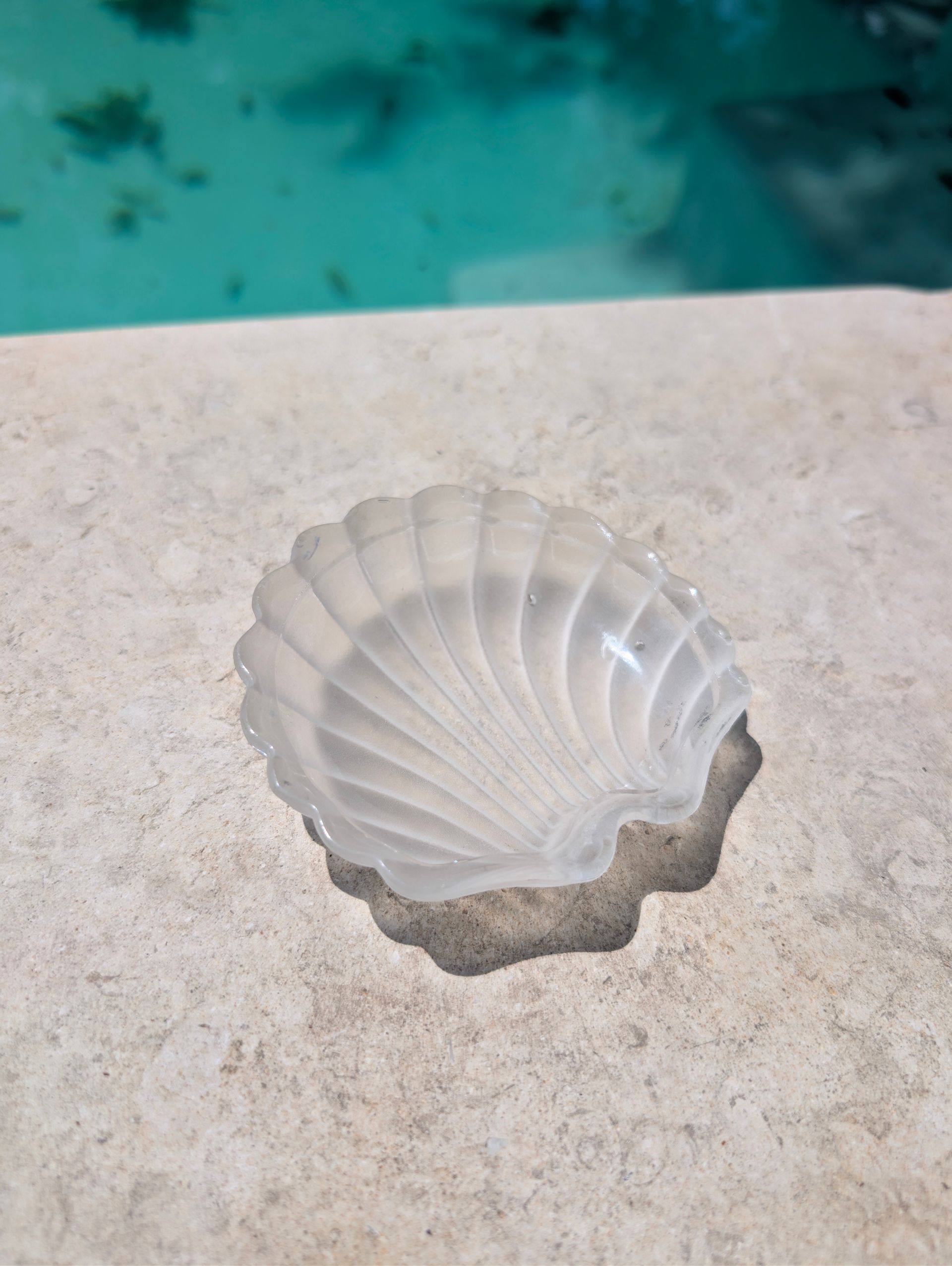 White scallop shell candle holder on a beige stone ledge by a turquoise pool.