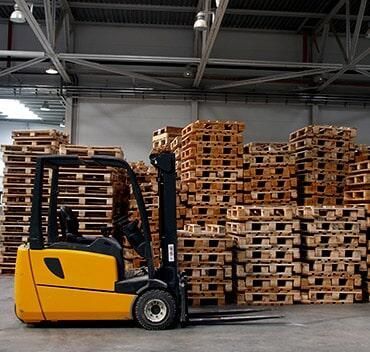 Una carretilla elevadora amarilla está estacionada frente a pilas de paletas de madera.