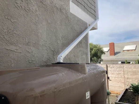 Rainwater harvesting system: white gutter draining into a brown tank next to a stucco wall.
