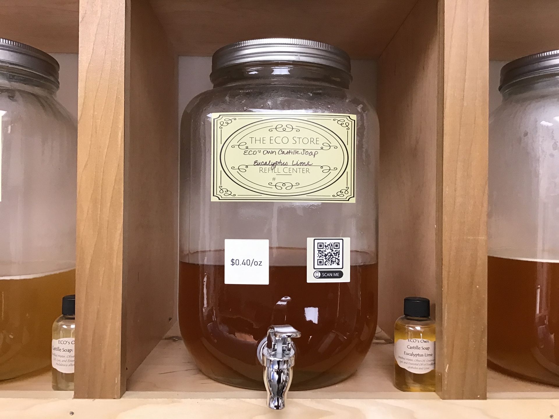 Large glass dispenser filled with brown liquid and labeled, on a wooden shelf, with small bottles in front.