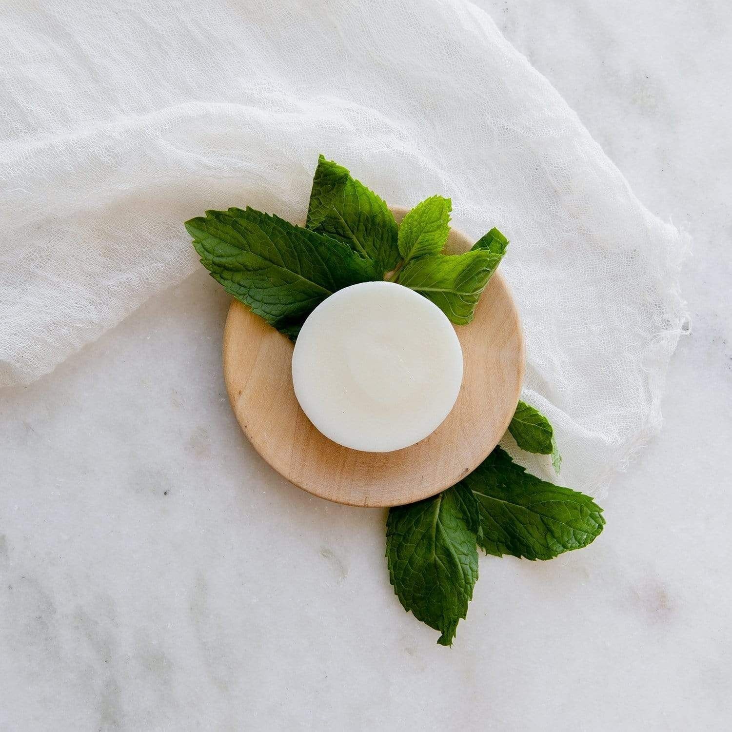 A white, round soap bar on a wooden dish, garnished with fresh mint leaves, against a white cloth backdrop.