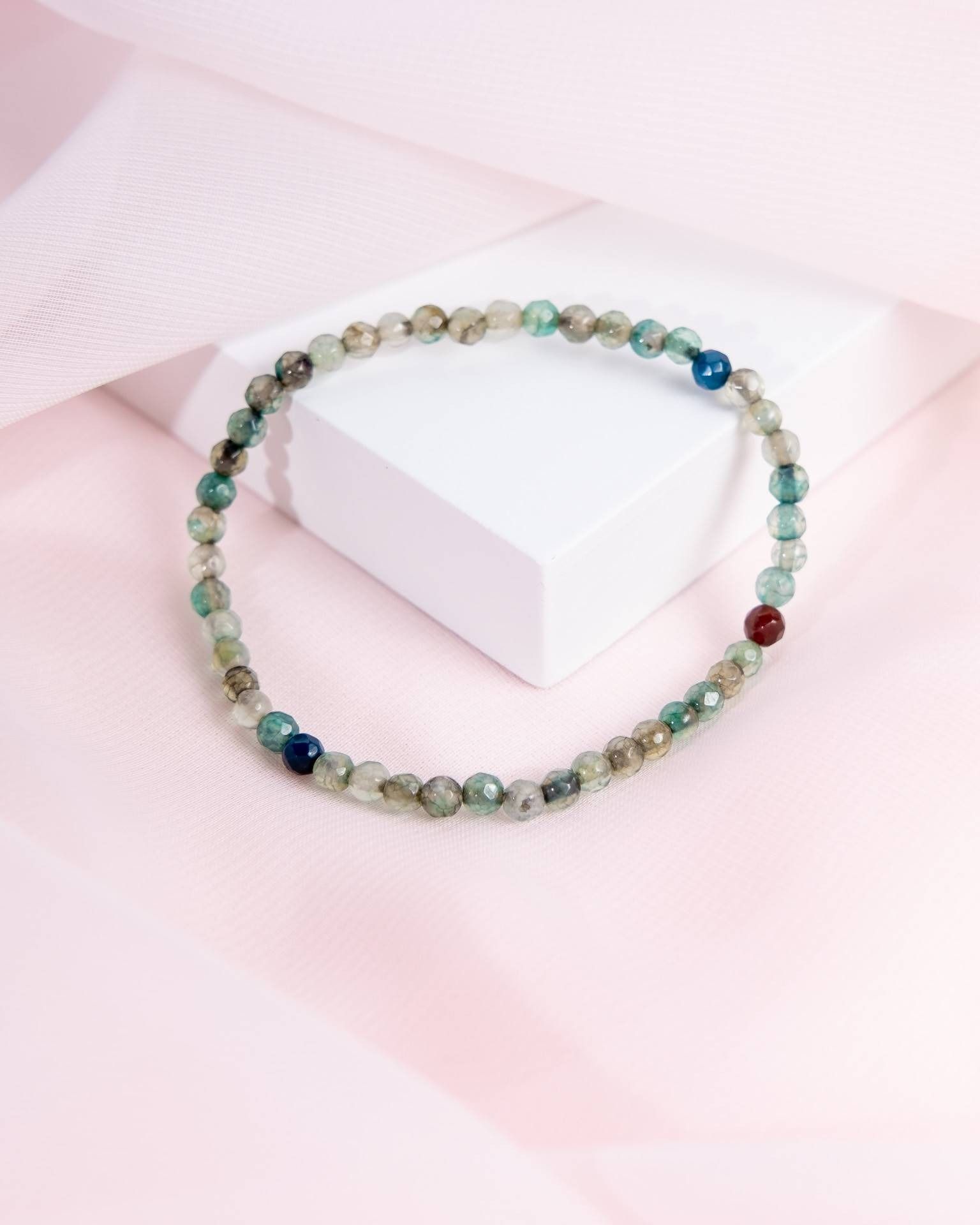 Beaded bracelet with faceted green and blue stones on a white block against a pink backdrop.