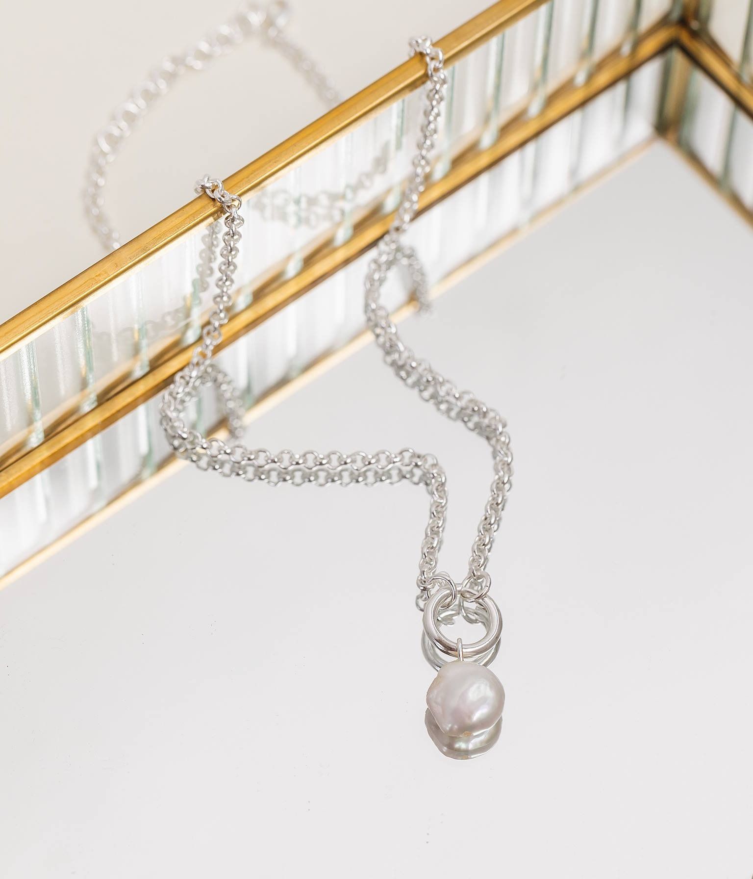 Silver necklace with a pearl pendant resting on a mirrored surface.