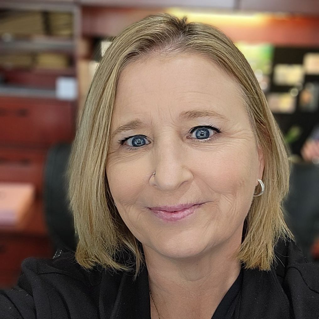 Blonde woman with blue eyes smiles at the camera, wearing a black jacket, indoors.