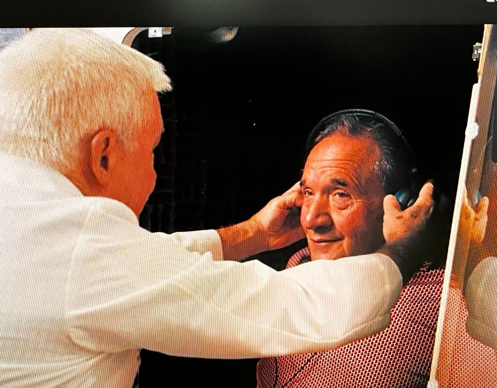Two men, one white-haired, adjusting headphones on the other's head. The other man smiles. Indoors, dark background.