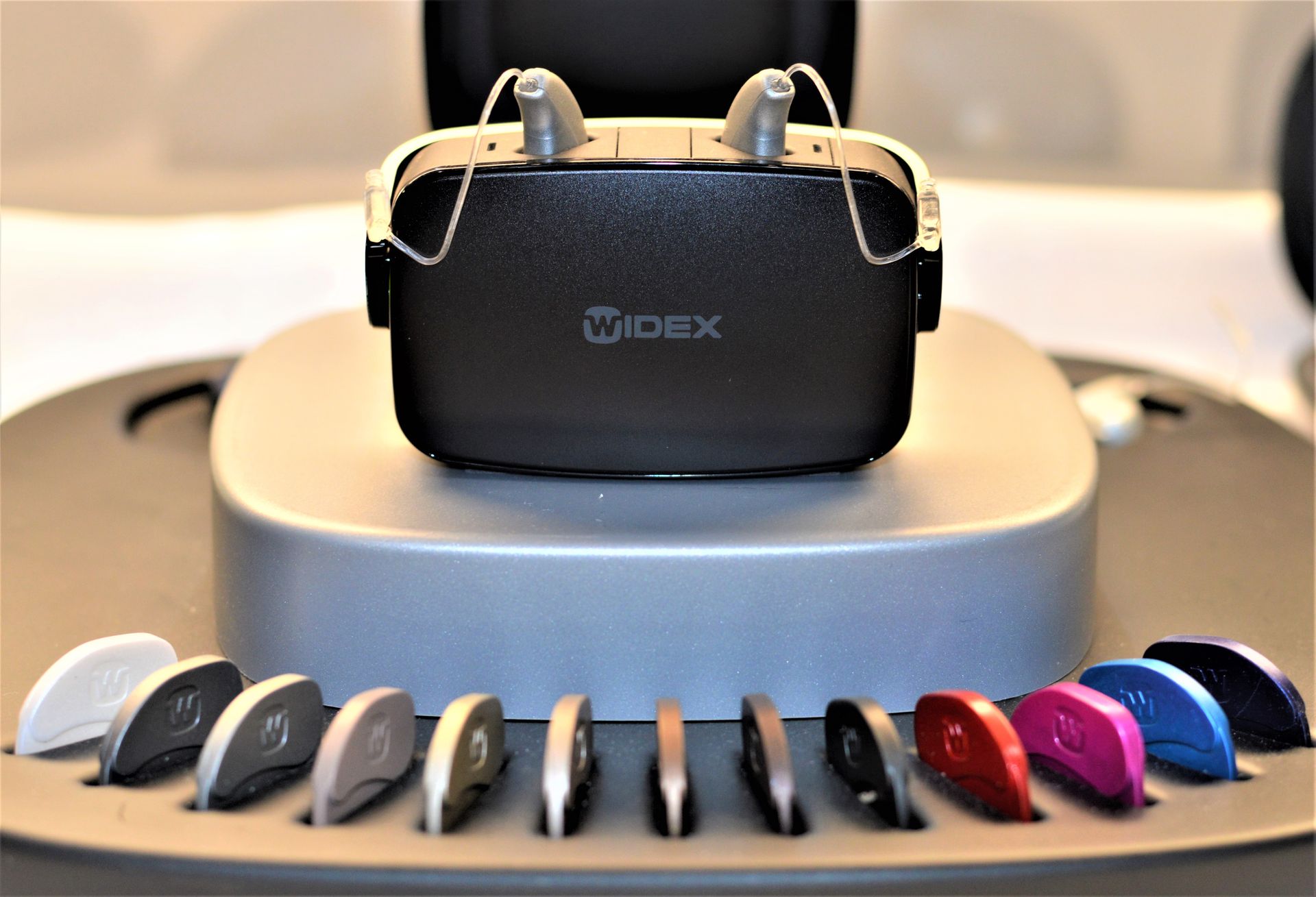 A black Widex charging case with hearing aids, displayed above a row of colored ear dome replacements.