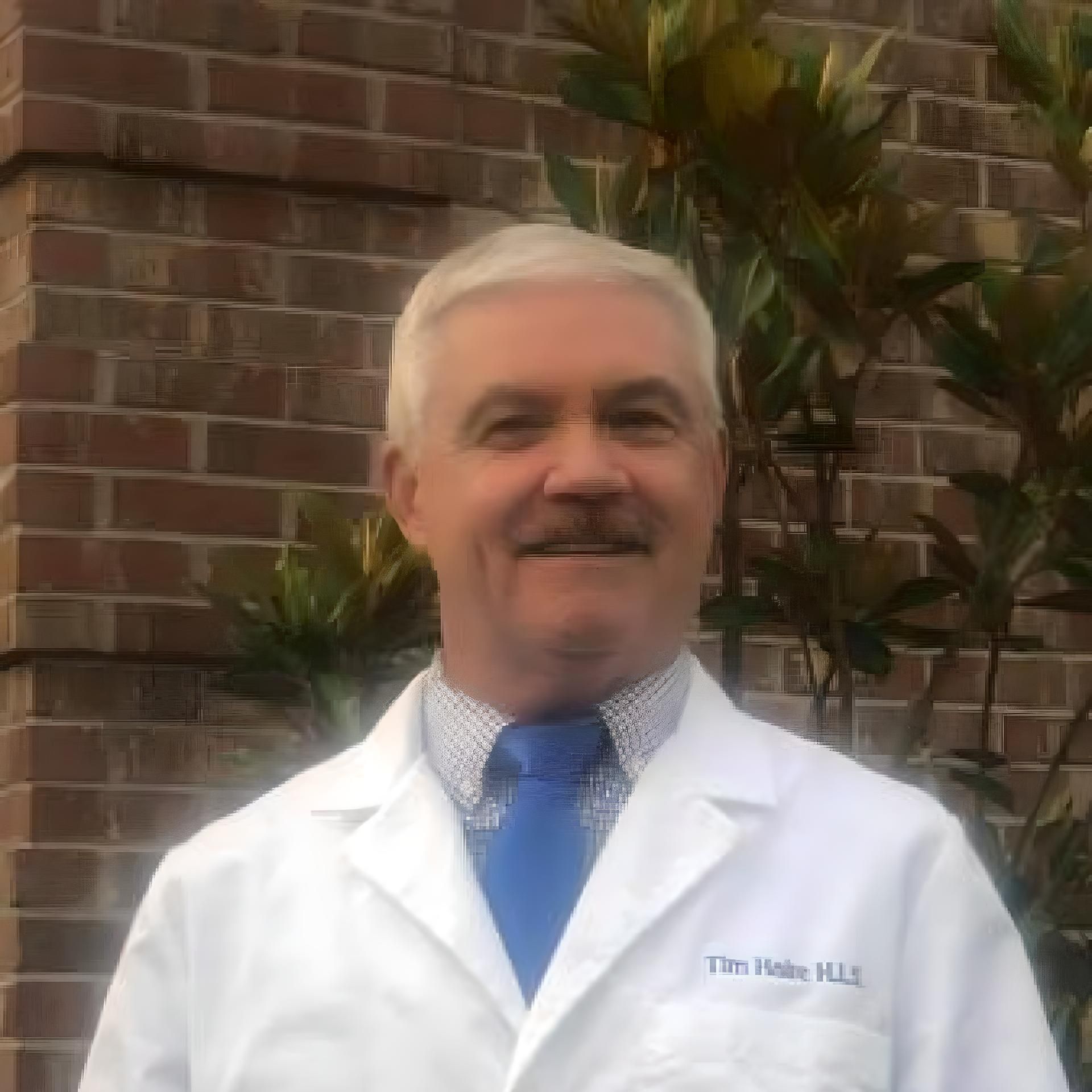 Man in white coat and blue tie smiles in front of a brick wall and green foliage. Name tag: 