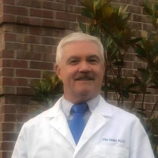Man in white coat and blue tie smiles in front of a brick wall and green foliage. Name tag: