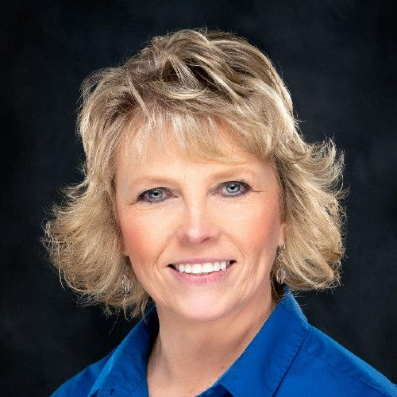 Woman with short blonde hair, blue eyes, and a warm smile, wearing a blue shirt against a dark background.