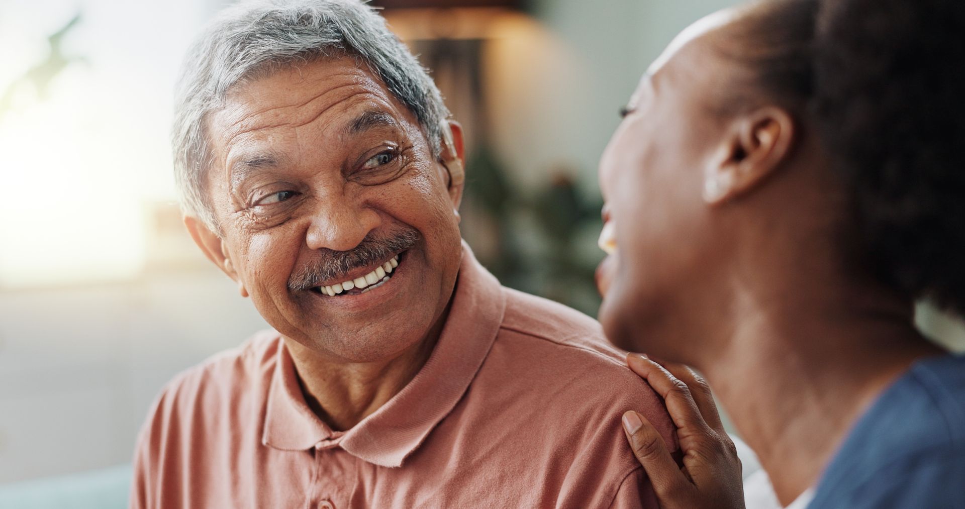 Smiling elderly Black man looks at a woman who has her hand on his shoulder. They are indoors.