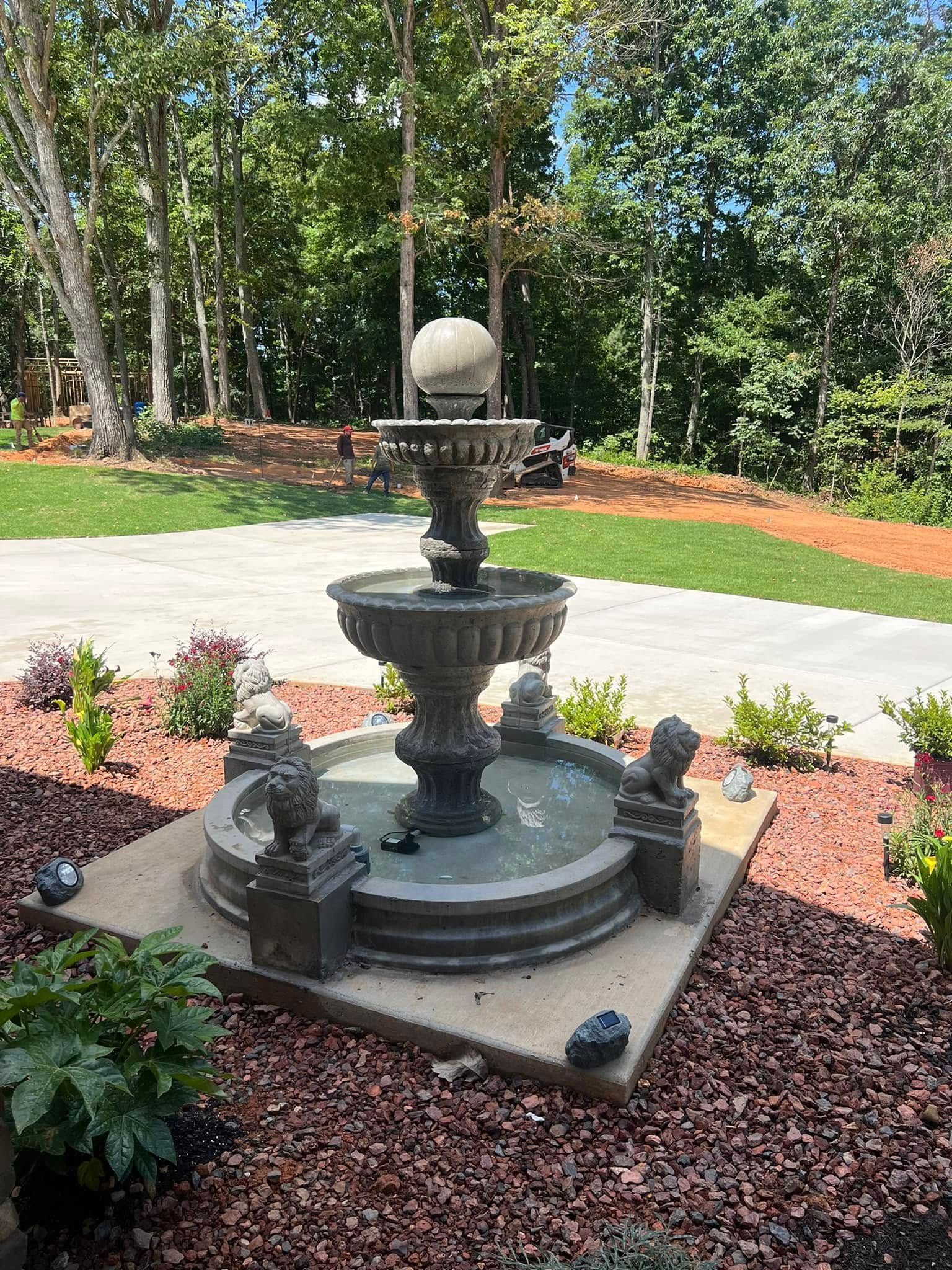 Decorative tiered garden fountain with stone lions, set in a landscaped yard with red mulch and trees