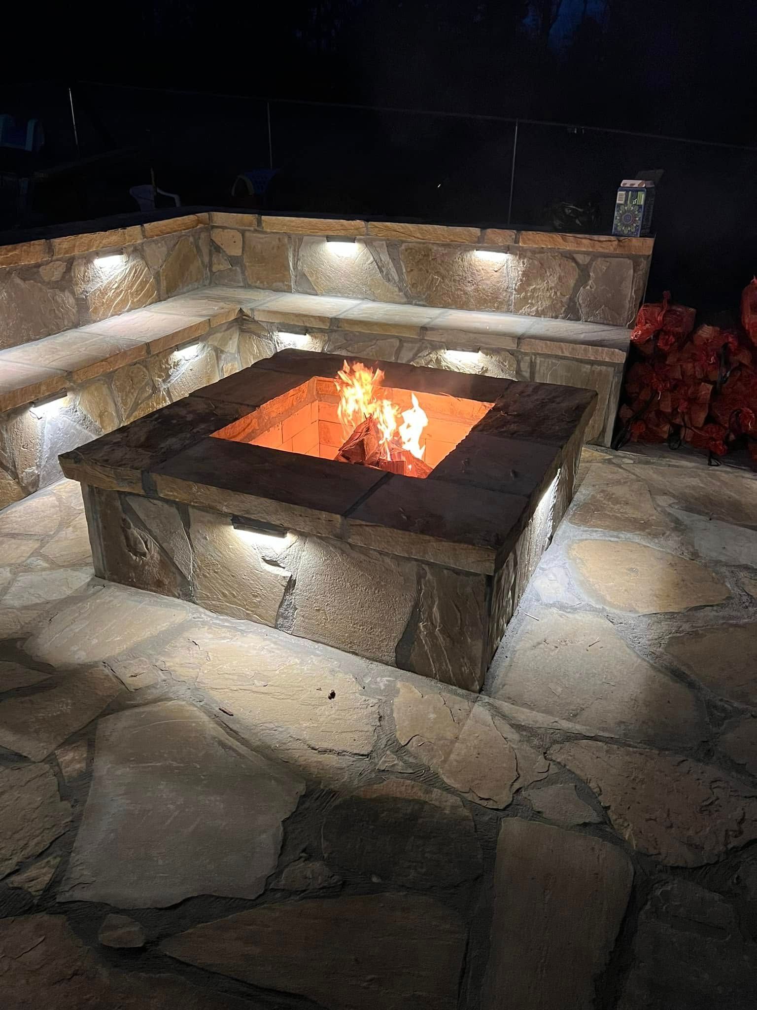 Stone patio fire pit with glowing flames at night, surrounded by built-in stone seating.