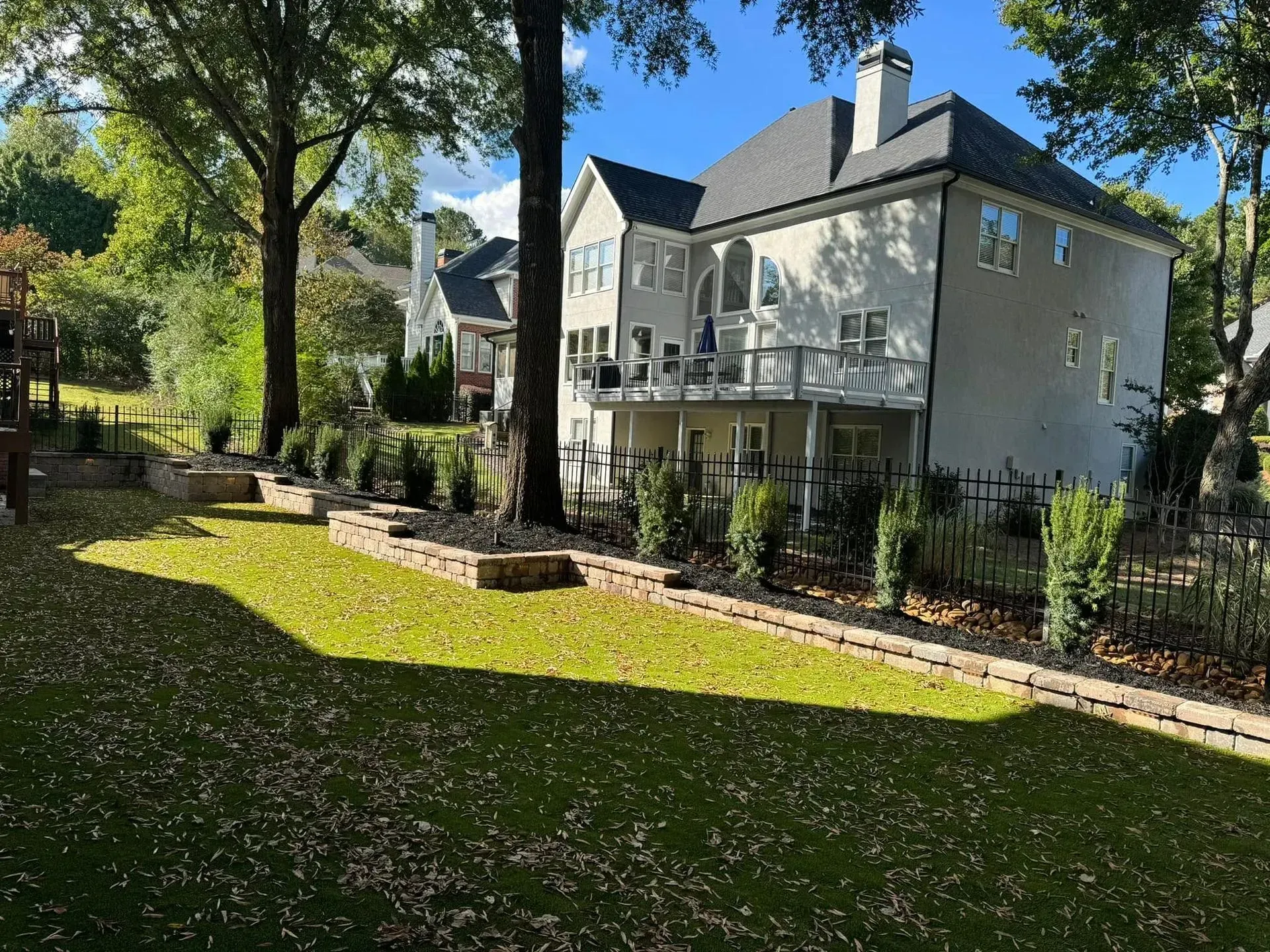Sunny backyard with a two-story house, patio, and large trees beside a grassy lawn