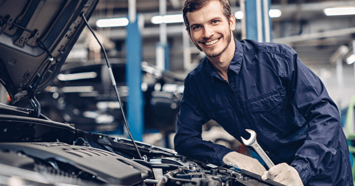 a smiling mechanics holding a wrench in front of a car