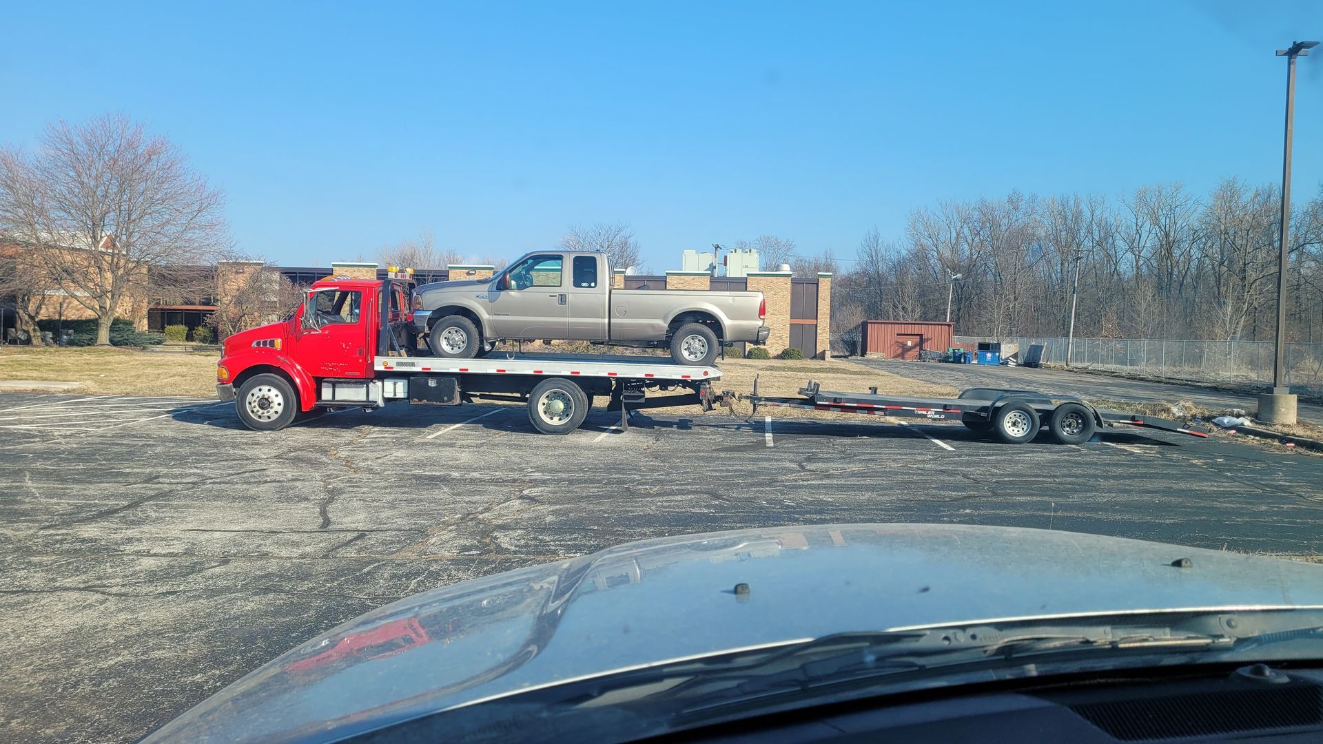 Red tow truck transporting a silver pickup truck on a flatbed trailer, parked in a lot.