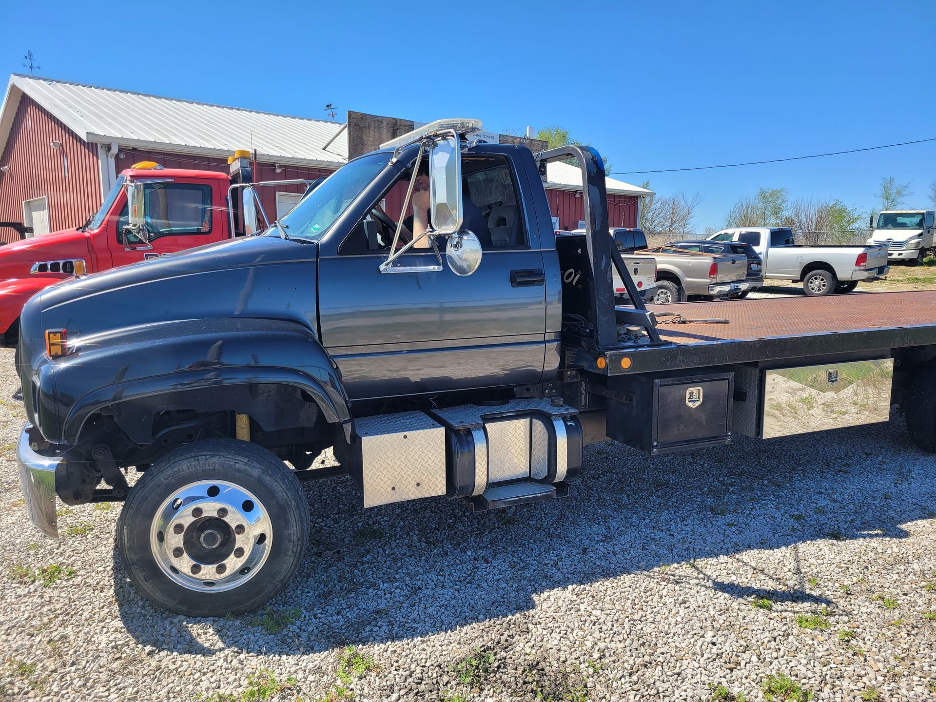 Black flatbed truck parked on gravel under a blue sky. Red truck in the background.